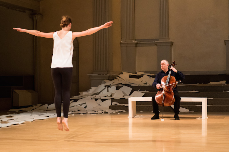 Butler leaps in the air with her back to the audience and her arms outstreched, Martin watches her as he plays the cello on the bench